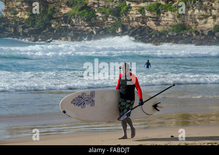 An older man carrying a standup paddleboard (SUP) along Freshwater Beach on a sunny Sydney morning in Australia Stock Photo