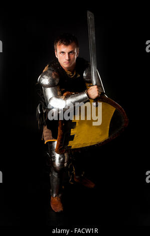 Studio shot of young man dressed as medieval knight armed with sword and shield in defense position over black background Stock Photo