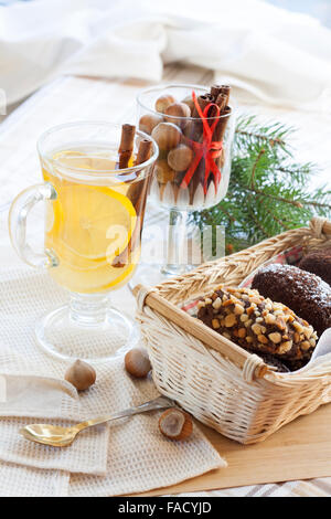 Fresh hot green tea in glass cup with lemon and cinnamon, chocolate cakes in basket and nuts and cinnamon in glass goblet. Stock Photo