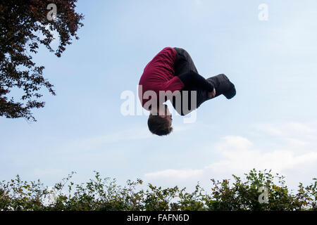 Millennial man on a trampoline upside down doing a forward roll somersault in mid air above a hedge against the blue sky. England, UK, Britain Stock Photo