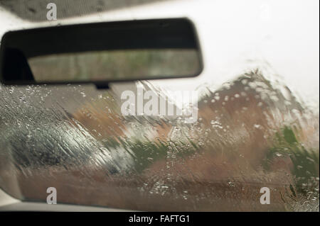 rear mirror and heavy down pour of rain on outside of car windscreen looking out to row house roof tops obscured water smear Stock Photo