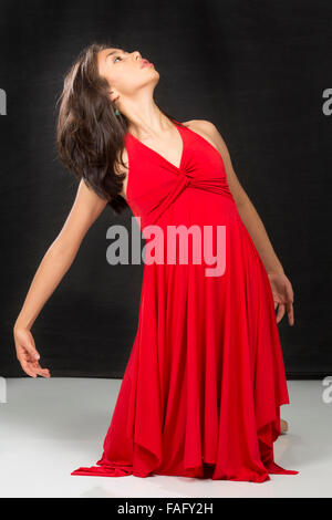 Attractive young woman dancer in red dress, leaning left with head in profile, arms down while kneeling, in floor movement. Stock Photo