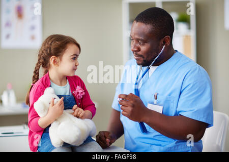 Cute girl with teddybear looking at clinician at hospital Stock Photo