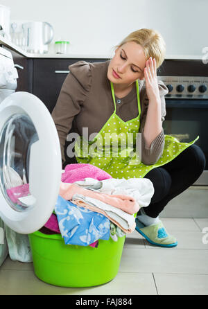 Tired blonde woman doing laundry with washing machine at home kitchen Stock Photo