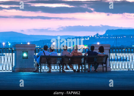 Friends sitting together, rear view of a group of middle aged friends sitting on a bench watching the afterglow of a colourful sunset in Sicily. Stock Photo