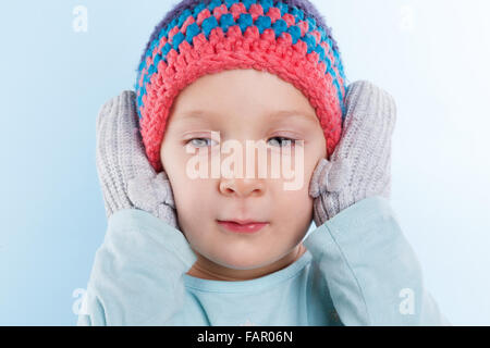 Cute baby girl in gloves covering her ears and hat isolated on light blue background. Winter season. Stock Photo