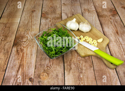 Fresh arugula leaves in a glass bowl, two bulbs of garlic, chopped garlic and a knife on an old wooden worktop on old wooden bro Stock Photo