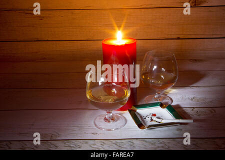 Two glasses of cognac, burning candles and burning cigarette in the ashtray in a romantic light on the background of a wooden ta Stock Photo