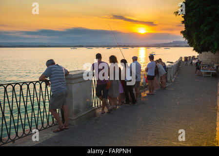 People sunset, view on a summer evening of people gathered on the esplanade in the harbour at Syracuse (Siracusa) to watch a colourful sunset, Sicily. Stock Photo