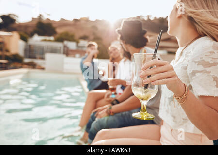Woman holding a cocktail glass while sitting on the edge of swimming pool with friends. Young people enjoying a poolside party w Stock Photo