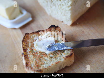 Spreading butter on a slice of toast with a butter dish and homemade loaf of bread behind Stock Photo