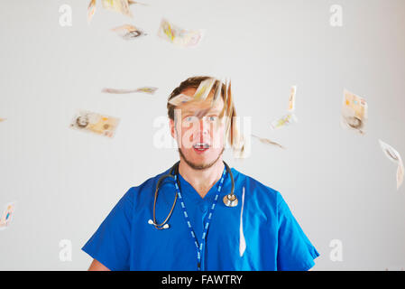 Young white male junior hospital doctor in theatre blues wearing a stethoscope throws a handful of ten pound notes into the air Stock Photo