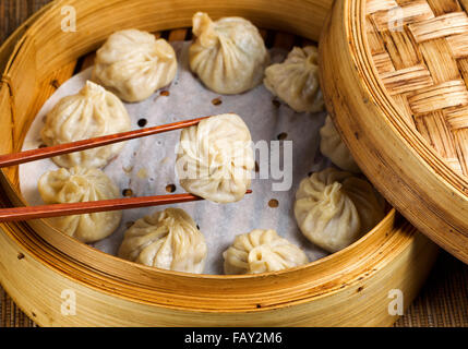 Close up of Chinese steamed dumplings, selective focus on piece held in chopsticks, being taken out of bamboo steamer Stock Photo