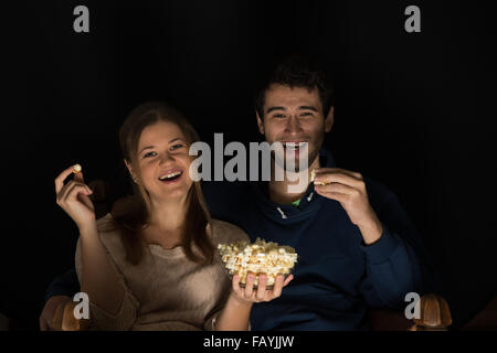 Young couple, man and woman, sitting in the dark room in the front of tv watching movie and eating popcorn, showing emotions Stock Photo
