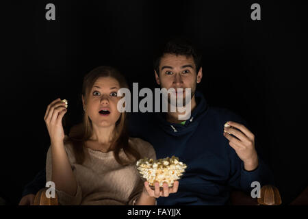 Young couple, man and woman, sitting in the dark room in the front of tv watching movie and eating popcorn, showing emotions Stock Photo
