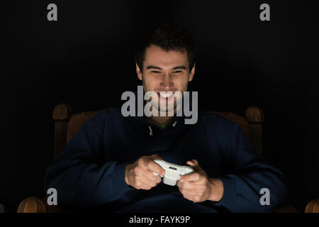 Young man plays computer games sitting in dark room in front of tv or monitor, holding game controller in his hands Stock Photo