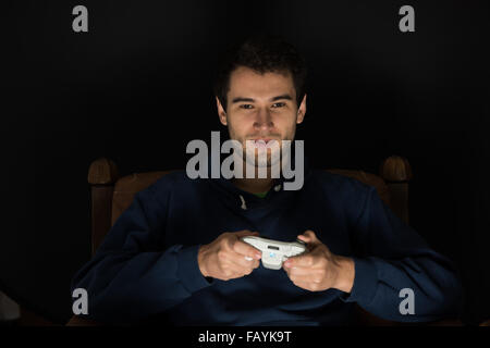 Young man plays computer games sitting in dark room in front of tv or monitor, holding game controller in his hands Stock Photo