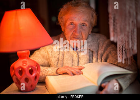 An old woman reads a book under a table lamp at night. Stock Photo
