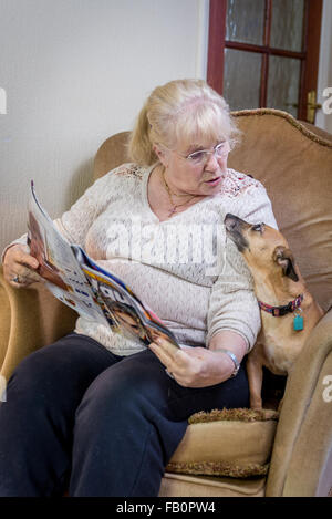 An Elderly lady at home reading a magazine and talking to her dog Stock Photo