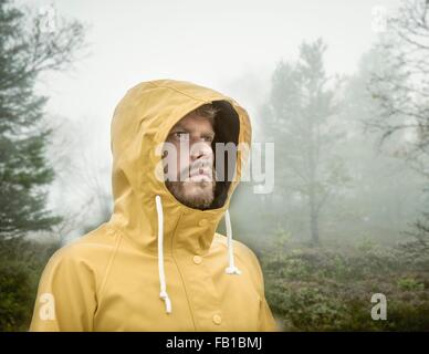 Bearded young man in misty forest wearing hooded yellow raincoat looking away Stock Photo