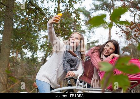 Teenage girls in forest sitting on bicycles using smartphone to take selfie smiling Stock Photo