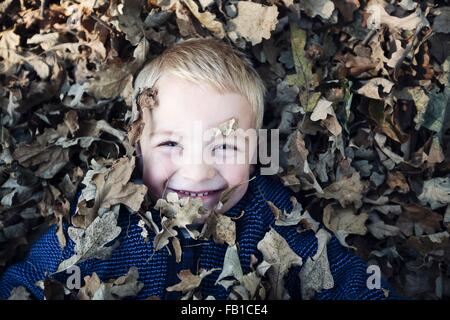 Overhead view of boy lying down covered in autumn leaves looking at camera smiling Stock Photo