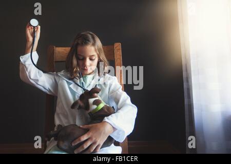 Girl sitting on chair dressed up as doctor tending to Boston terrier puppy using stethoscope Stock Photo