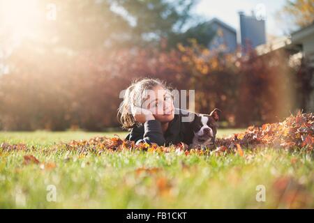 Girl with Boston terrier puppy lying on front on Autumn leaf covered grass, resting on elbow looking at camera Stock Photo