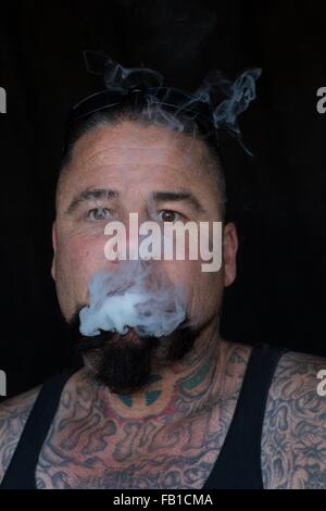 Portrait of bearded tattooed mature man looking at camera blowing smoke Stock Photo