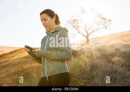 Female runner on sunlit hill choosing smartphone music Stock Photo