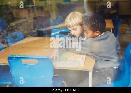 Window view of two boys doing puzzle at desk in elementary school classroom Stock Photo