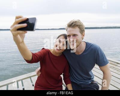 Couple on pier side by side using smartphone to take selfie smiling, Copenhagen, Denmark Stock Photo