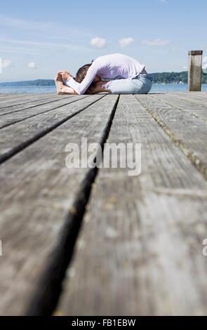 Surface level side view of mature woman sitting on pier bending over touching toes Stock Photo