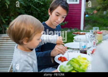 Teenage boy helping female toddler eat food at garden barbecue Stock Photo
