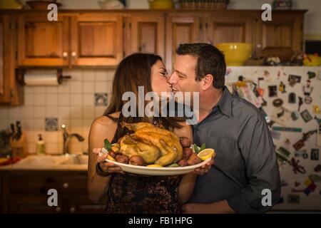 Mature couple kissing whilst holding roast chicken dinner in kitchen Stock Photo
