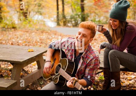 Young couple playing guitar on picnic bench in autumn forest Stock Photo