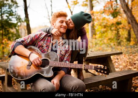 Romantic young couple playing guitar on picnic bench in autumn forest Stock Photo