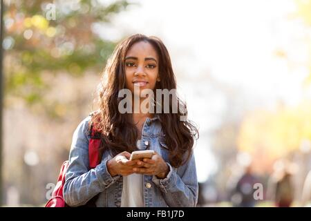 Young woman with long brown hair wearing denim jacket holding smartphone looking away smiling