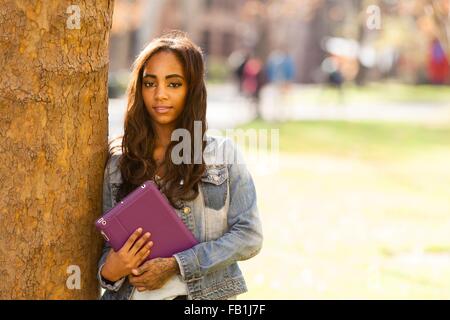 Portrait of young woman leaning against tree holding digital tablet looking at camera smiling Stock Photo