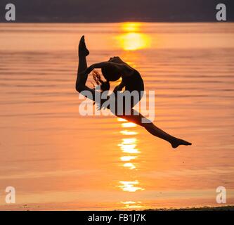 Side view of girl in silhouette by ocean at sunset in mid air, legs apart throwing head back Stock Photo