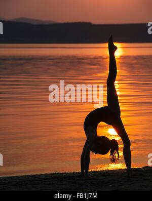 Side view of girl in silhouette by ocean at sunset bending over backwards leg raised doing the splits Stock Photo