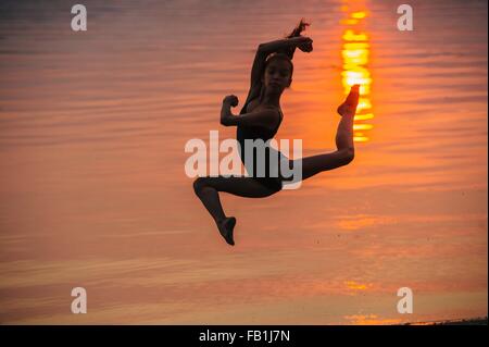Side view of girl in silhouette by ocean at sunset leaping in mid air, legs apart looking at camera Stock Photo