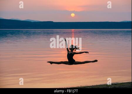 Side view of girl by ocean at sunset leaping in mid air, arms raised doing the splits Stock Photo