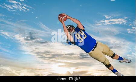 Male teenage American football player catching ball mid air against blue sky Stock Photo