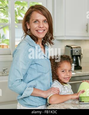 Side view of mother behind daughter in kitchen looking at camera smiling Stock Photo