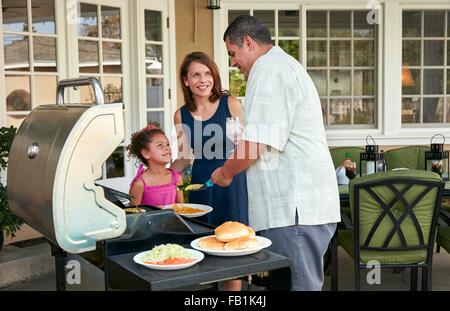 Mother and girl on patio holding plate being served barbeque food by father Stock Photo