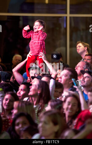Perched above the multiracial crowd on her father's shoulders, an excited toddler watches a stage show at a Christmas festival in a San Clemente, CA, shopping mall. Stock Photo