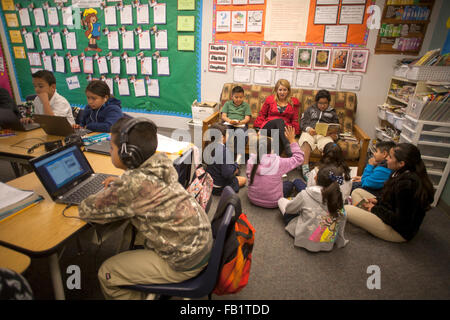 An elementary school teacher holds a reading seminar for her Hispanic students in the corner of her classroom in San Clemente, CA. Note posters describing reading goals, book nook at right, and students using Google Chromebook laptops in foreground. Stock Photo