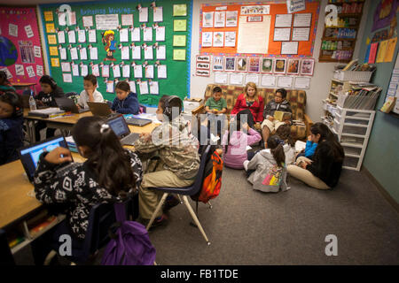 An elementary school teacher holds a reading seminar for her Hispanic students in the corner of her classroom in San Clemente, CA. Note posters describing reading goals, book nook at right, and students using Google Chromebook laptops in foreground. Stock Photo