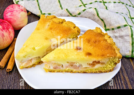 Two pieces of apple pie with cream sauce in white plate, cinnamon, apples and a napkin on a wooden boards background Stock Photo